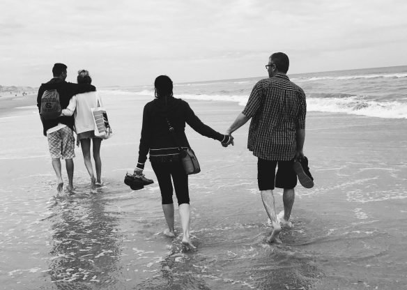 Two couples holding hands and walking on a sandy beach with gentle waves.
